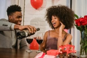 Positive young black couple in elegant clothes sitting at table while man pouring red wine from bottle in glasses near gifts and vase with bouquet with roses at light home